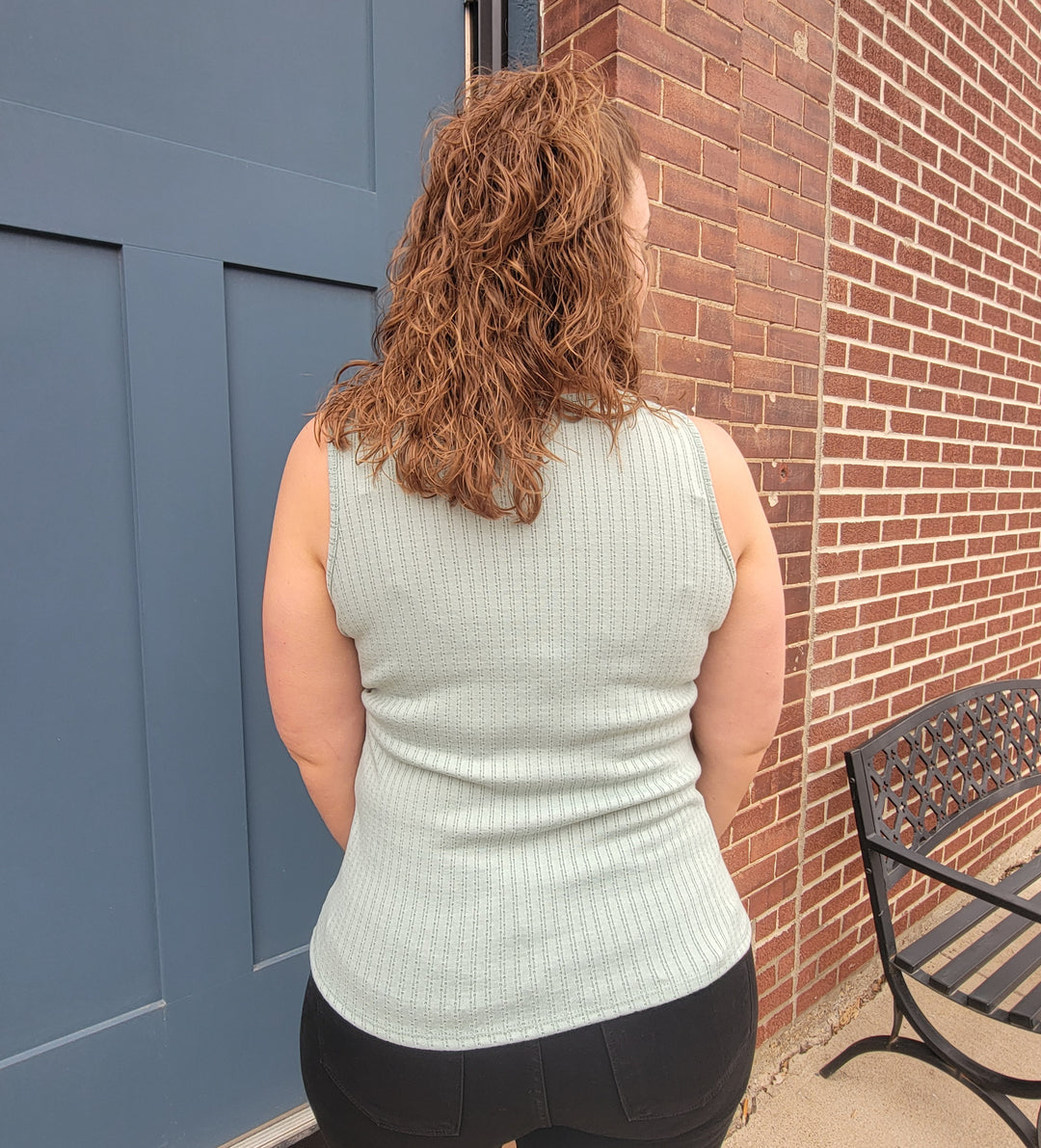 A person with curly, shoulder-length brown hair is seen from behind in front of a blue door and brick wall, wearing the 7th Ray Palmer Button Front Tank Top in light blue and black pants, next to a metal bench.