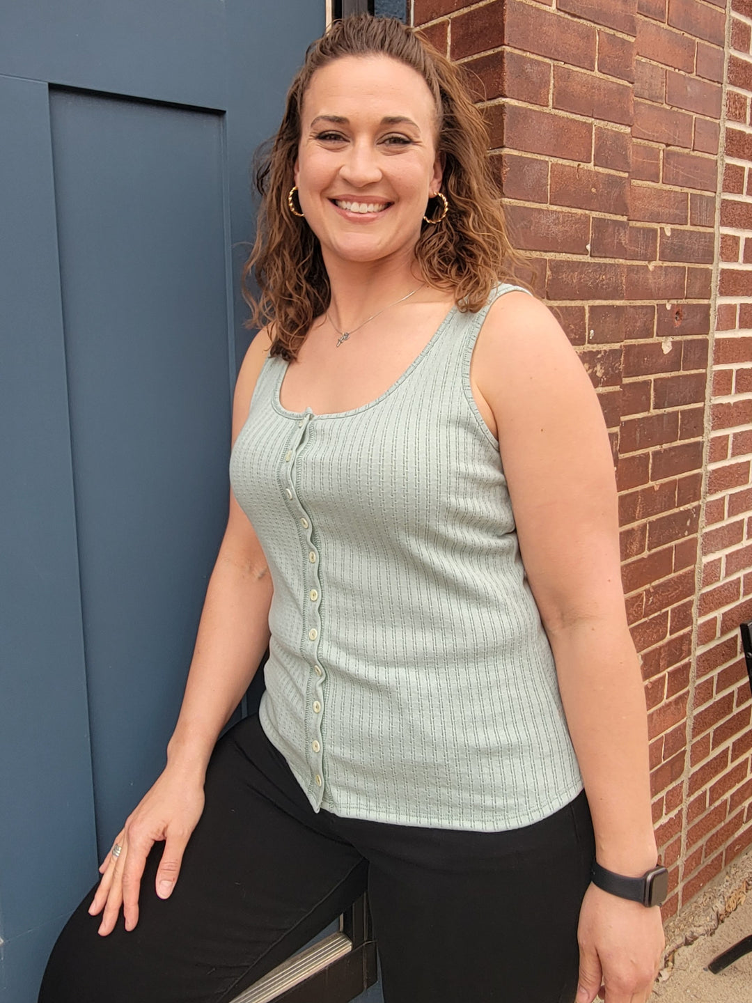 A smiling woman with wavy brown hair stands by a blue door and brick wall, wearing the 7th Ray Palmer Button Front Tank Top in light green, along with black pants, hoop earrings, and a smartwatch.