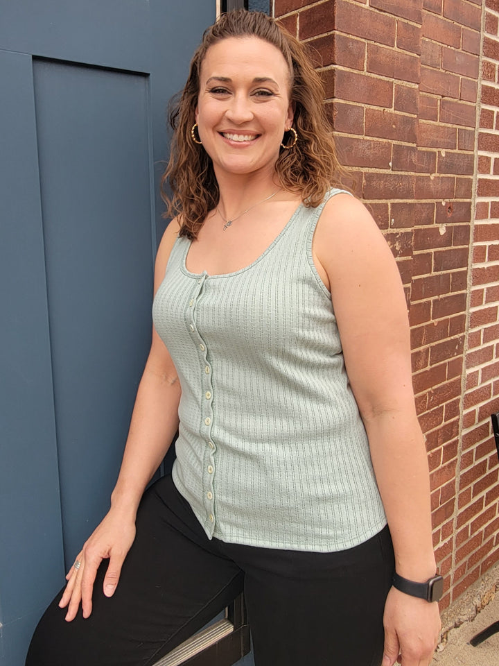 A smiling woman with wavy brown hair stands by a blue door and brick wall, wearing the 7th Ray Palmer Button Front Tank Top in light green, along with black pants, hoop earrings, and a smartwatch.
