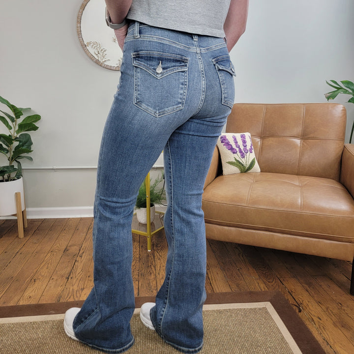 Someone wearing Judy Blue's Brielle Flare Jean and a gray shirt stands on a rug in a living room featuring a tan leather chair, plant-themed pillow, indoor plants, and a round mirror on the wall.