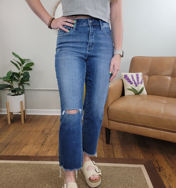 A person stands indoors on a rug wearing Lovervet’s Estella Straight Jean, featuring a ripped knee, paired with a gray crop top, cream sandals, and a watch. Behind them are a tan couch with a pillow and a potted plant.