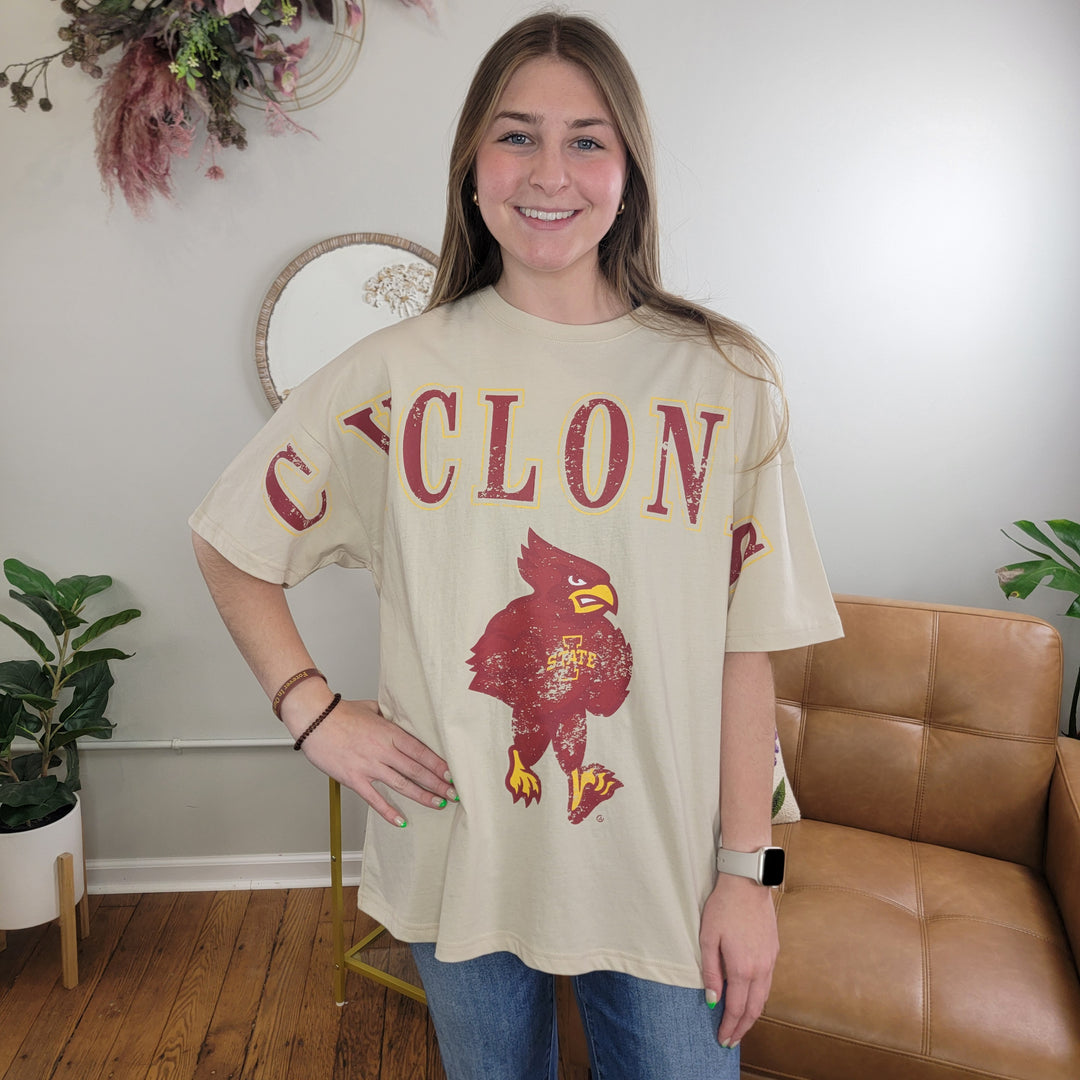 A smiling woman stands indoors in a cozy living room, wearing Game Day Social’s Cyclone's Tee—a beige Iowa State Cyclone shirt with CYCLONE and a red bird graphic—perfect attire for cheering on game day.