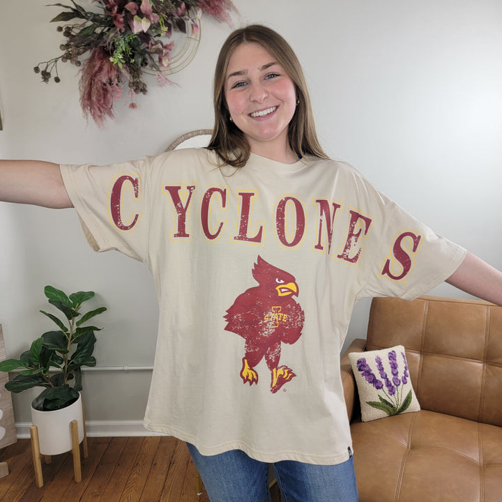 A smiling woman stands indoors with arms outstretched, wearing the Game Day Social Cyclone's Tee—a beige Iowa State t-shirt with a red and yellow bird mascot—near a tan couch, houseplants, and pink dried flower wall art.