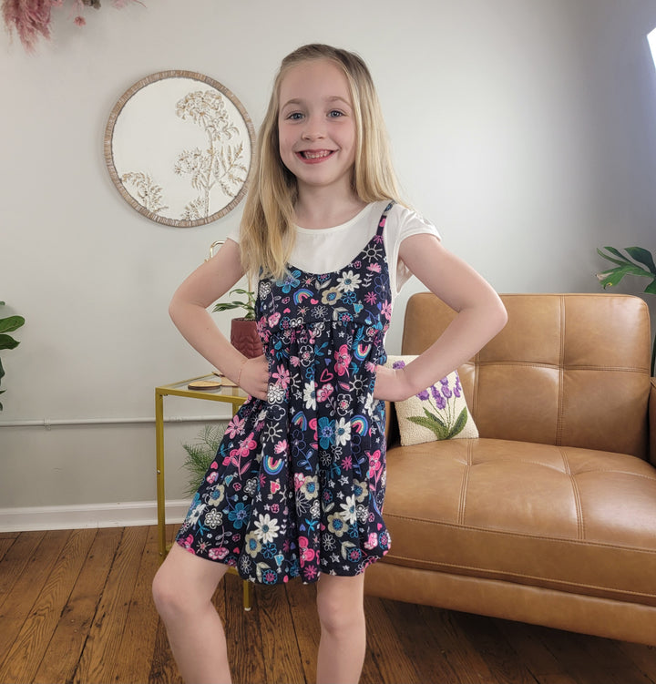 A young girl with long blonde hair smiles confidently, hands on hips, wearing the Girls Georgia Dress by Cutie Patootie. Behind her are a brown leather sofa, yellow side table, and a decorative round wall mirror.