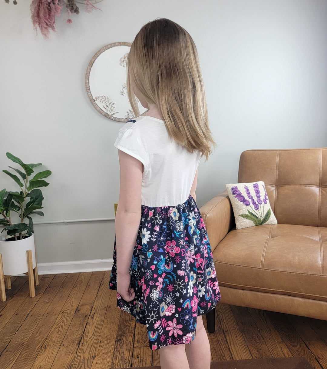 A young girl with shoulder-length light brown hair stands indoors facing away, wearing the Cutie Patootie Girls Georgia Dress. Around her are plants, a tan chair with a lavender pillow, and a round mirror on the wall.