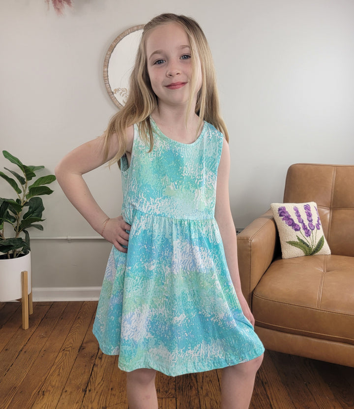 A young girl with long blonde hair smiles in the Cutie Patootie Kids: Fenella Dress II, a sleeveless cotton dress. She poses by a tan couch and plant in a bright living room with hardwood floors and light decor.