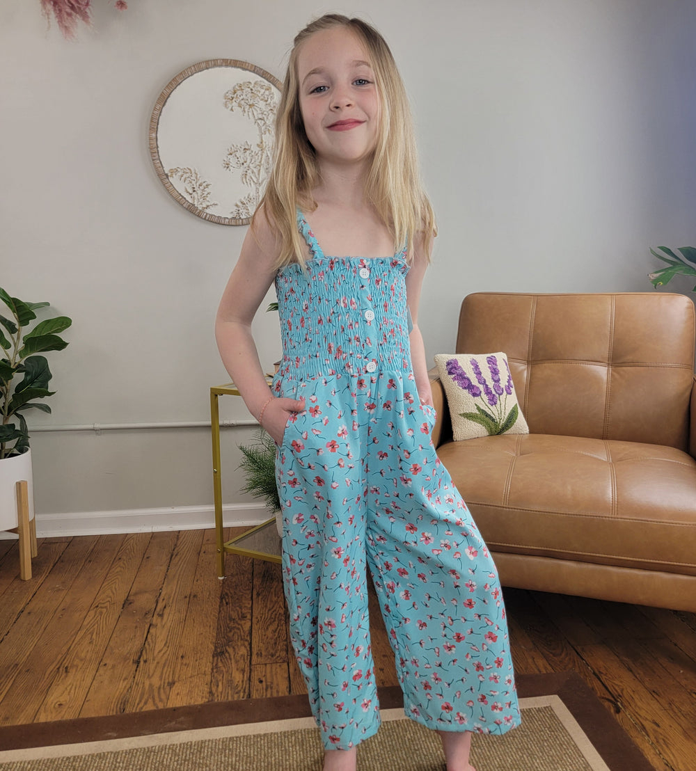 A young girl with long blonde hair smiles indoors, wearing the Cutie Patootie Kids: Melody Jumper II—a light blue jumper with a pink flower print. She stands in a cozy room with wooden floors, a leather sofa, and plants.