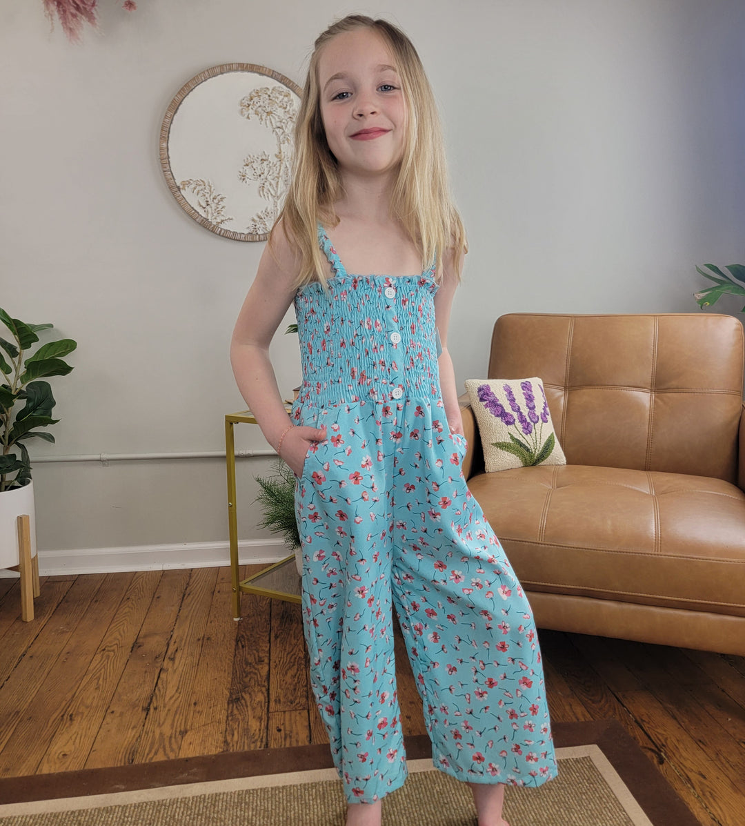 A young girl with long blonde hair smiles indoors, wearing the Cutie Patootie Kids: Melody Jumper II—a light blue jumper with a pink flower print. She stands in a cozy room with wooden floors, a leather sofa, and plants.