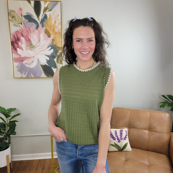 A smiling woman with wavy brown hair wears the Staccato Taini Olive Sleeveless Sweater made from 100% cotton and blue jeans, standing in a cozy living room with plants, a leather chair, and floral artwork on the wall.