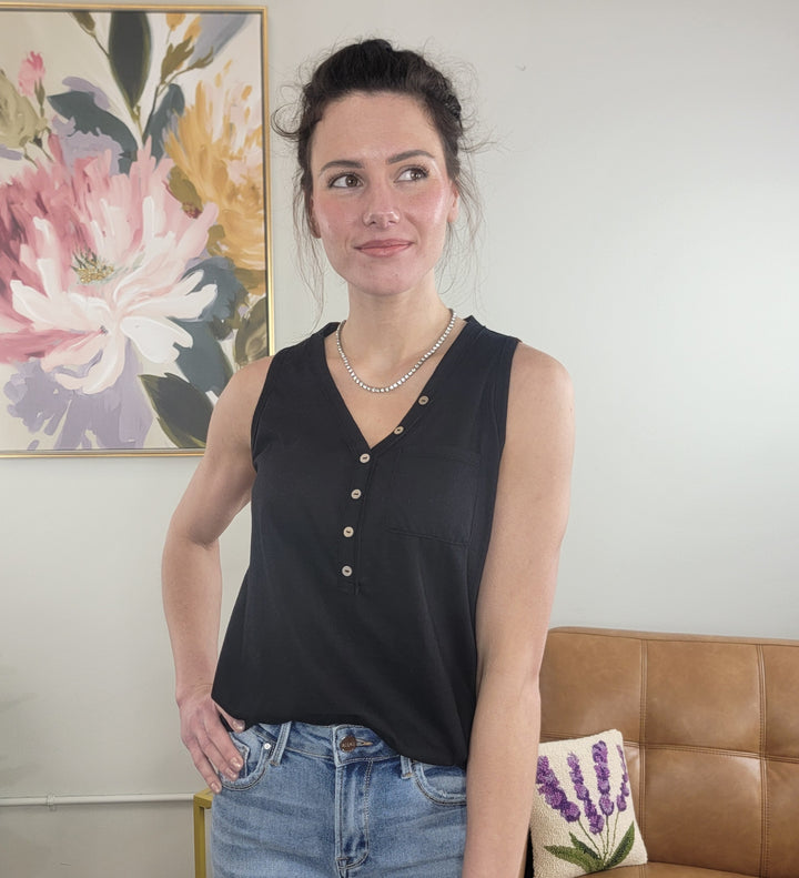 A woman with dark hair in a messy bun wears the HYFVE, Inc. Myra Tank—a lightweight black v-neck top—and blue jeans. She stands indoors, smiling slightly in front of a tan couch and floral painting, looking off to the side.