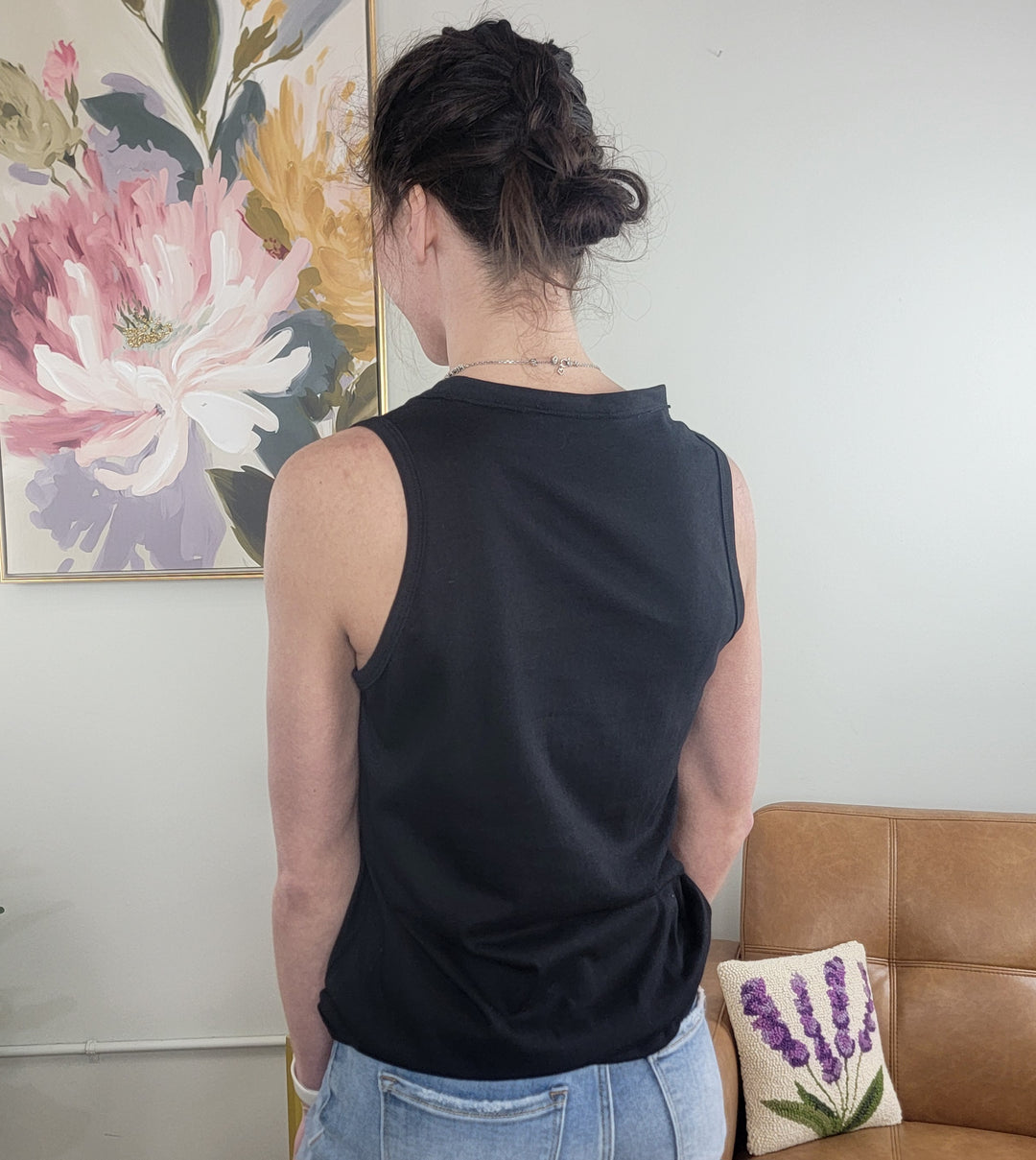 A person with dark hair in a bun wears the HYFVE, Inc. Myra Tank and jeans, standing with their back to the camera. Nearby are a floral painting and a small pillow featuring a lavender design on a tan couch.
