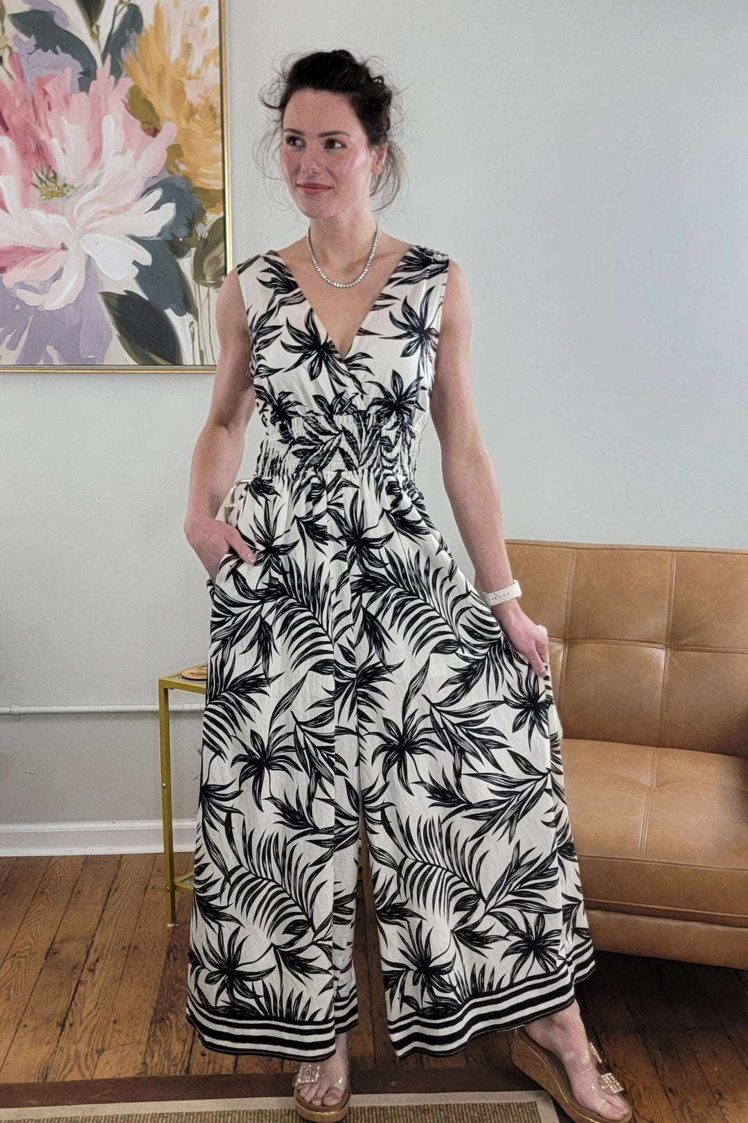 Indoors, a woman wears the Staccato Jadin Jumpsuit—a black-and-white v-neck summer style—paired with clear heeled sandals, pearl jewelry, and an updo. A floral painting and tan couch appear in the background.