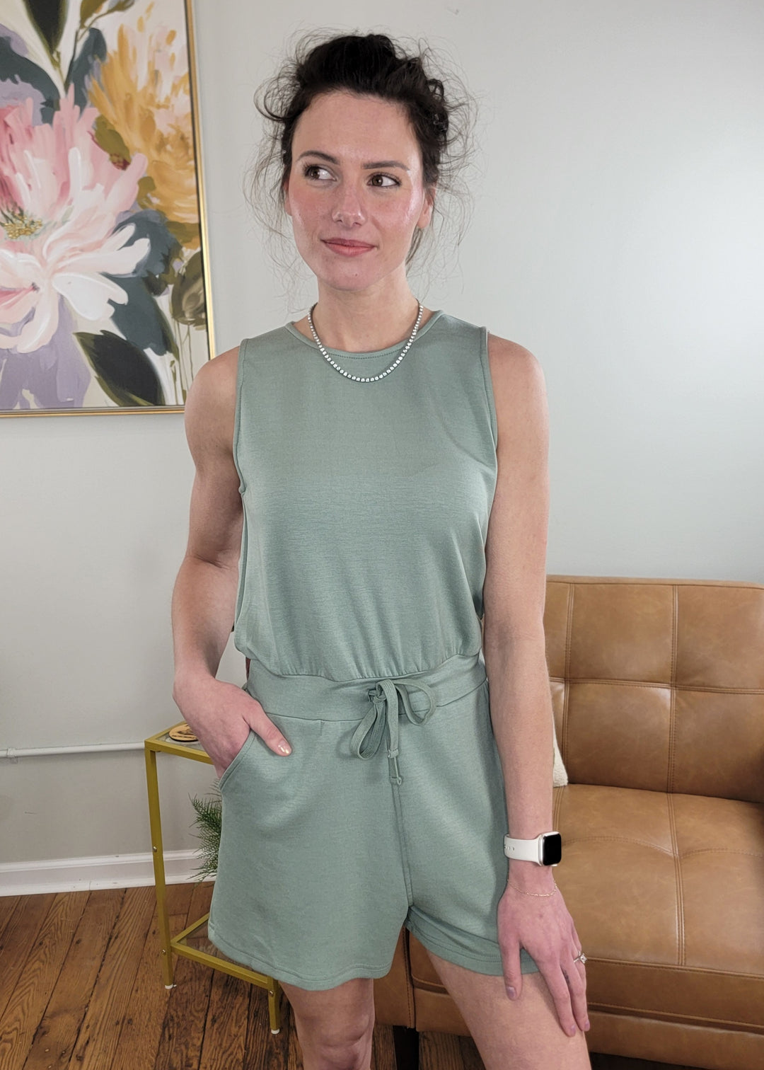 A woman wears the sage green Marsha Romper by HYFVE, Inc., indoors with one hand in her pocket, accessorized with a pearl necklace and Apple Watch, standing before a tan couch and floral wall art.