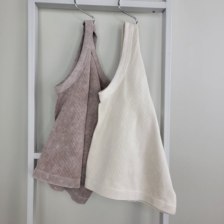 Two Superline Laverne Ribbed Tank tops—one taupe, one off-white—hang on a white rack against a pale wall, showcasing their slim fit and sleeveless design, ideal for summer layering.