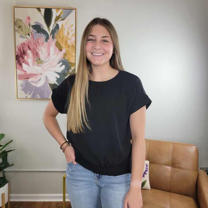 A woman with long brown hair smiles in a living room, wearing the Staccato Sutton Black Blouse with a wide crew neck and light blue jeans. Behind her are a tan chair, green plant, and a large floral painting on the wall.