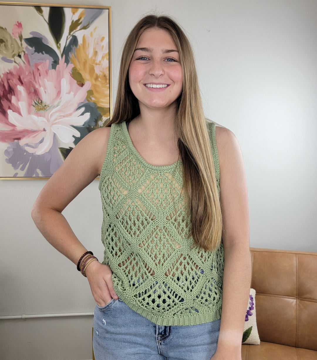 A smiling young woman with long hair wears the Robyn Open Knit Tank by HYFVE, Inc.—a sage green, diamond-patterned knit top—standing indoors before a beige couch and floral painting, with one hand in her jean pocket.