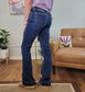 A person wearing Blue Element Bootcut Jeans from Flying Monkey stands on a wooden floor beside a tan sofa with a lavender pillow, a small yellow table, and potted plants in a cozy living room.