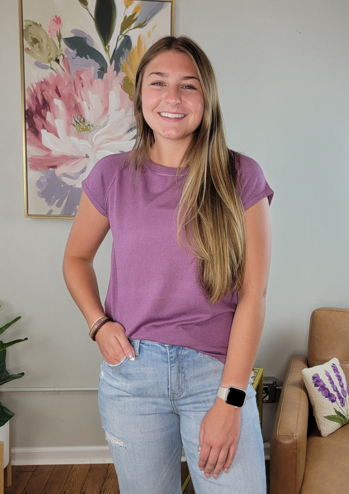 A young woman with long brown hair, wearing the Staccato Loxley Top in orchid and light blue jeans, stands smiling indoors. Behind her are a flower painting, tan chair, and plant on a wooden floor.