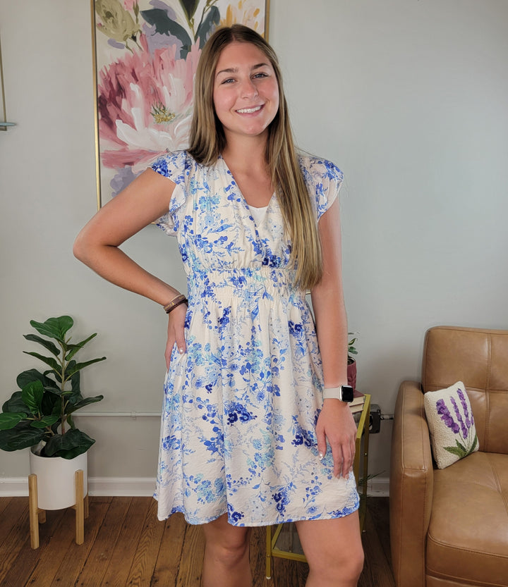 A woman with long brown hair smiles indoors wearing the Blakely Floral Dress by Staccato, featuring blue and white florals and flutter sleeves. She stands by a brown leather chair, green potted plant, and large floral painting.