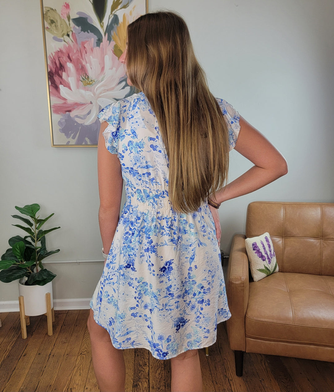 A woman with long hair stands indoors facing away, wearing the Staccato Blakely Floral Dress with flutter sleeves and a smocked waistband beside a tan chair, potted plant, and colorful floral artwork on the wall.