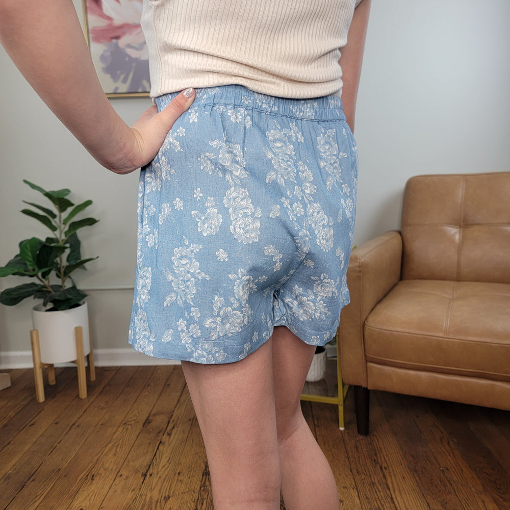 A person wearing the Hayden Girls' Janine Skort in light blue floral and a cream ribbed top stands indoors on a wooden floor near a tan leather chair and green potted plant, with one hand on their hip.