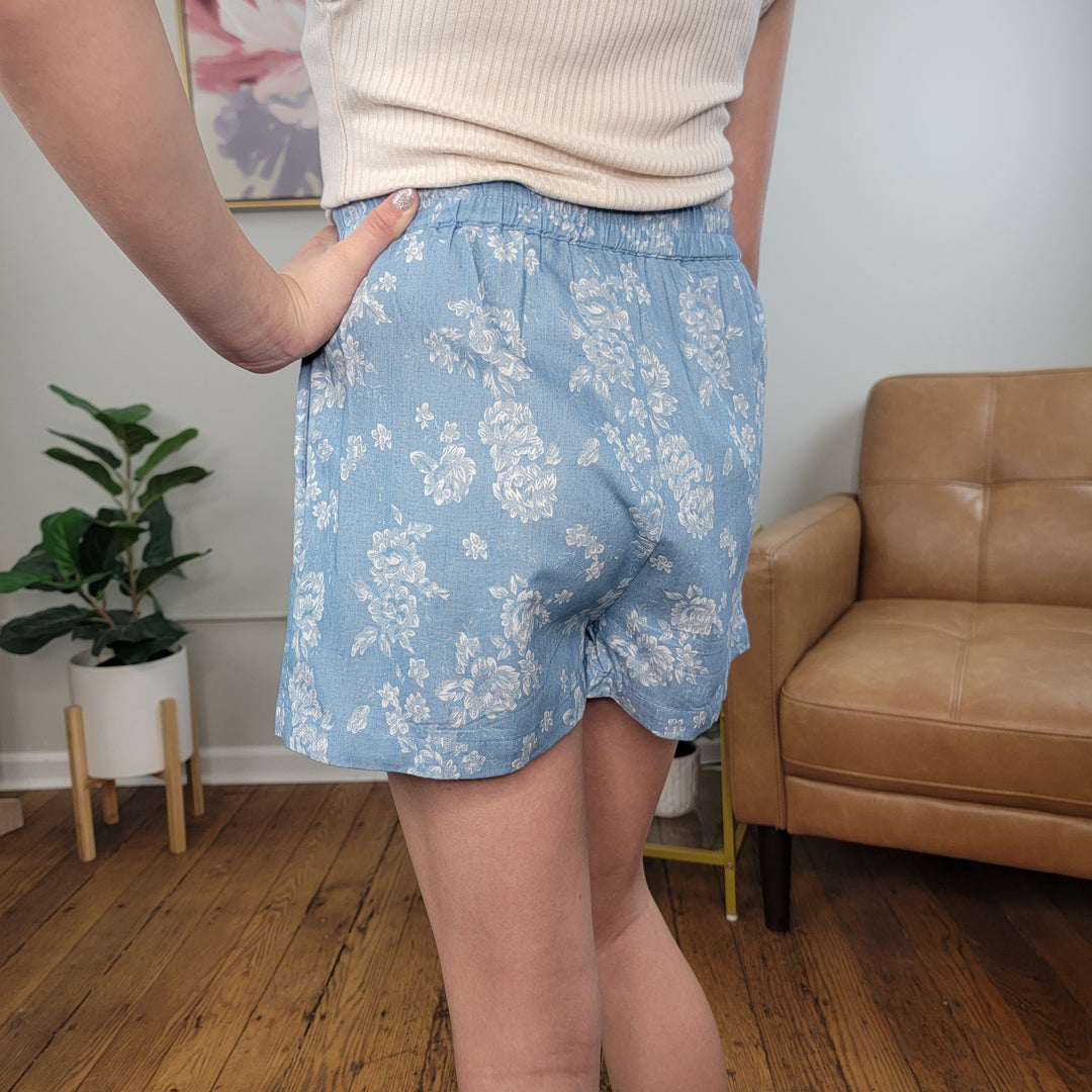A person wearing the Hayden Girls' Janine Skort in light blue floral and a cream ribbed top stands indoors on a wooden floor near a tan leather chair and green potted plant, with one hand on their hip.