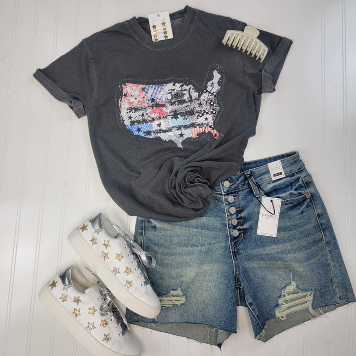 A flat lay of an outfit featuring Cosmos + Clover Boutique's Distressed USA Tee, distressed denim shorts, white star-patterned sneakers, gold star earrings, and a cream hair claw on a white wood background.