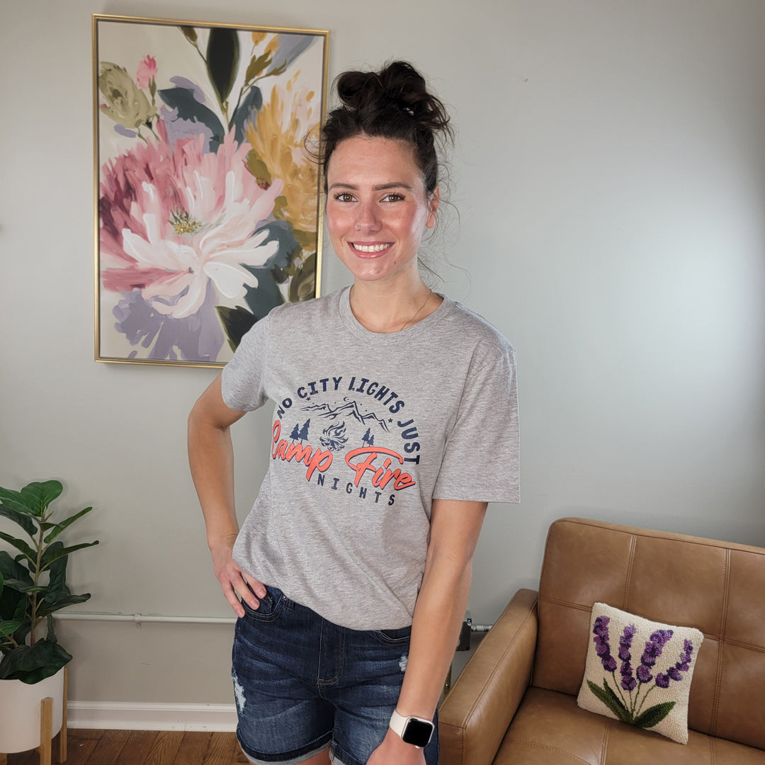 A woman with brown hair in a bun, wearing a gray Cosmos + Clover Boutique Campfire Graphic Tee and denim shorts, smiles in a living room decorated with floral accents and featuring a tan armchair.