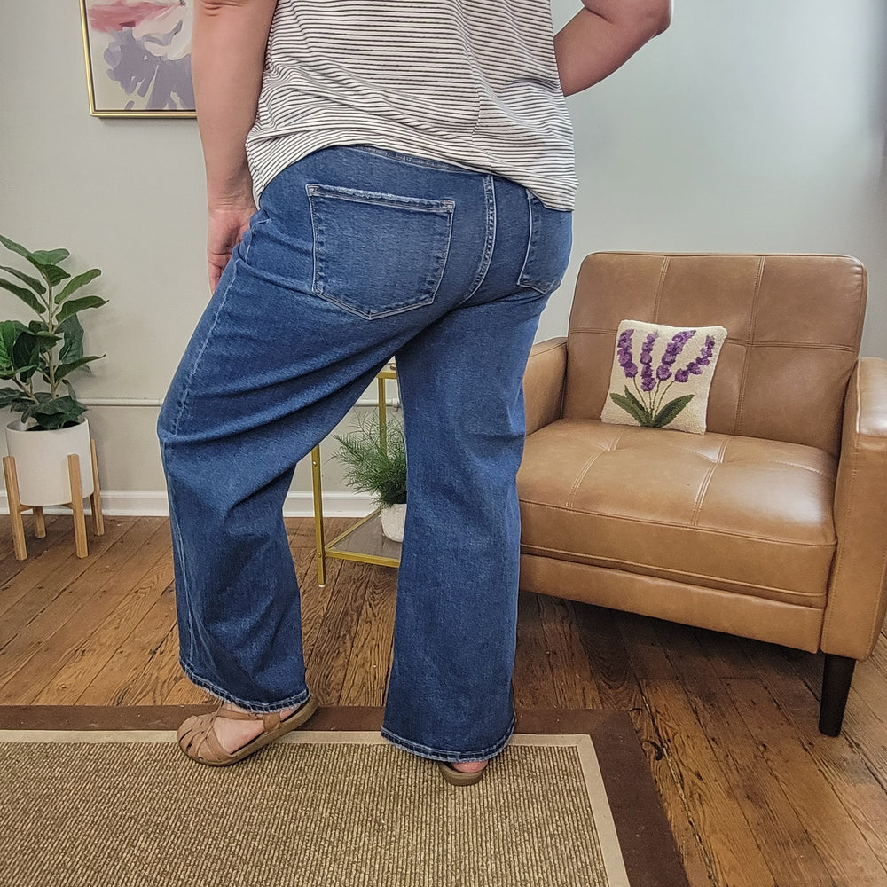 Wearing Risen's Emmet Wide Leg Crops, a striped shirt, and tan shoes, a person stands beside a tan leather chair in a cozy room featuring plants and wooden flooring.