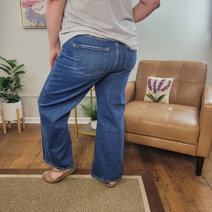 Wearing Risen's Emmet Wide Leg Crops, a striped shirt, and tan shoes, a person stands beside a tan leather chair in a cozy room featuring plants and wooden flooring.
