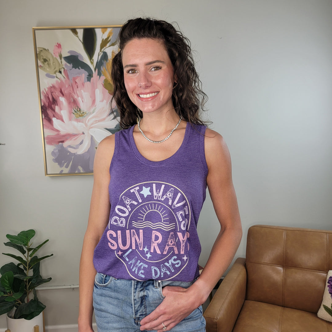 A woman with wavy brown hair smiles in a living room, wearing the Cosmos + Clover Boutique Boat Waves Graphic Tank—a purple racerback featuring the phrase "Boat Waves, Sun Rays & Lake Days"; floral art and a tan chair are in the background.