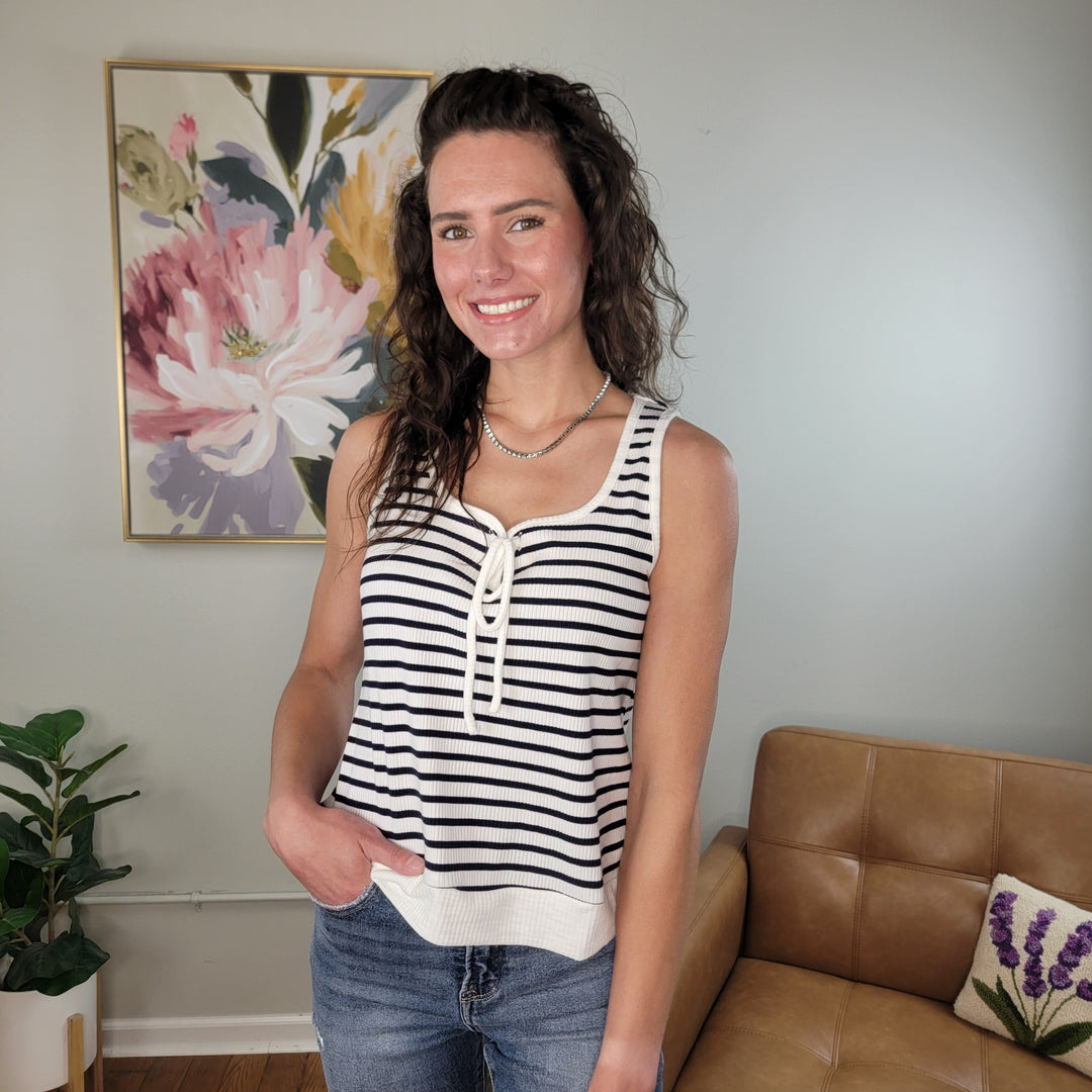 A woman with wavy brown hair smiles in a living room, wearing the Doe & Rae Shelby Tank—a black and white striped top—with jeans. Behind her are a floral painting, a potted plant, and a tan leather couch.