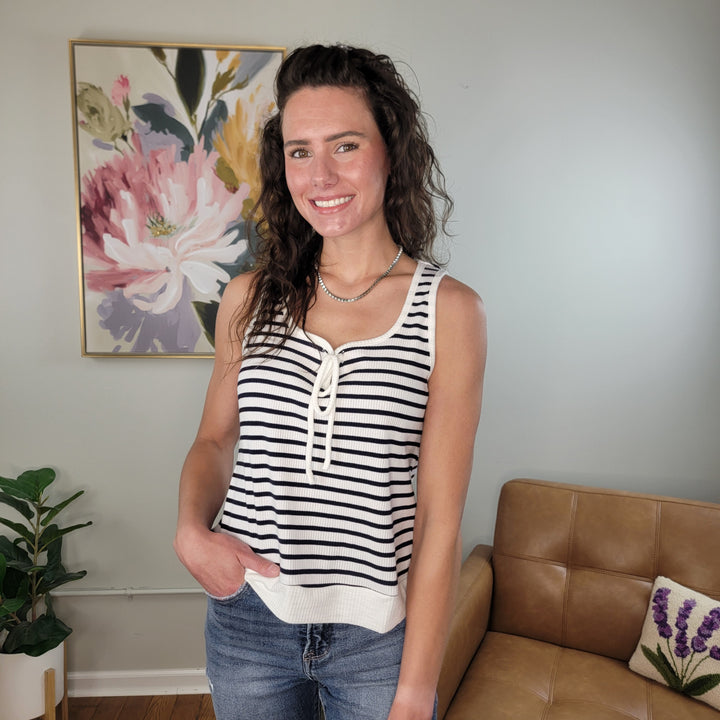 A woman with wavy brown hair smiles in a living room, wearing the Doe & Rae Shelby Tank—a black and white striped top—with jeans. Behind her are a floral painting, a potted plant, and a tan leather couch.