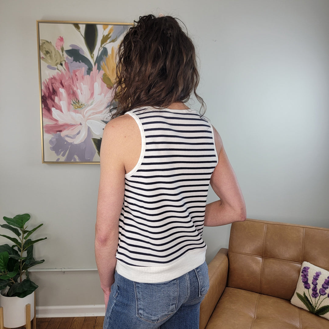 A person with wavy brown hair and blue jeans, seen from behind, wears the Doe & Rae Shelby Tank in a room with a floral painting, brown couch, and green plants.