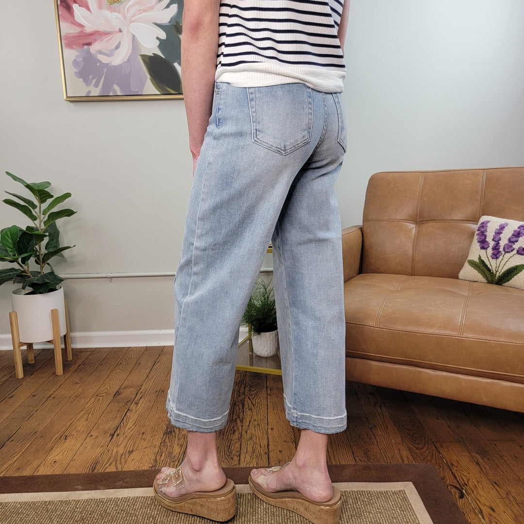A person stands indoors on a wooden floor wearing Risen’s Cassidy Crop Wide Leg Jeans with cuffed hems, a striped top, and clear platform sandals. The background features a tan sofa, green plants, and framed artwork.
