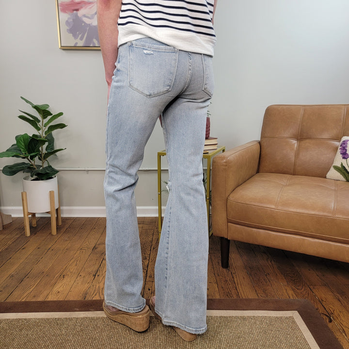 A person wearing the Risen Erica Flares stands on a wooden floor in a modern living room with tan shoes, a tan leather chair, a potted plant, and wall art.
