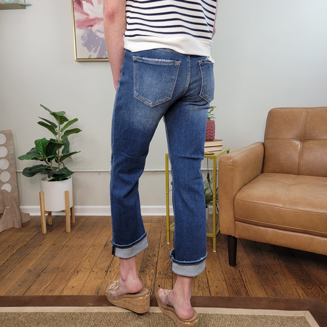 A person wearing Risen's Harlow Slim Straight Jeans, a striped top, and clear wedge sandals stands on a wooden floor by a tan armchair and small plant-topped table, with framed art and extra greenery in the room.