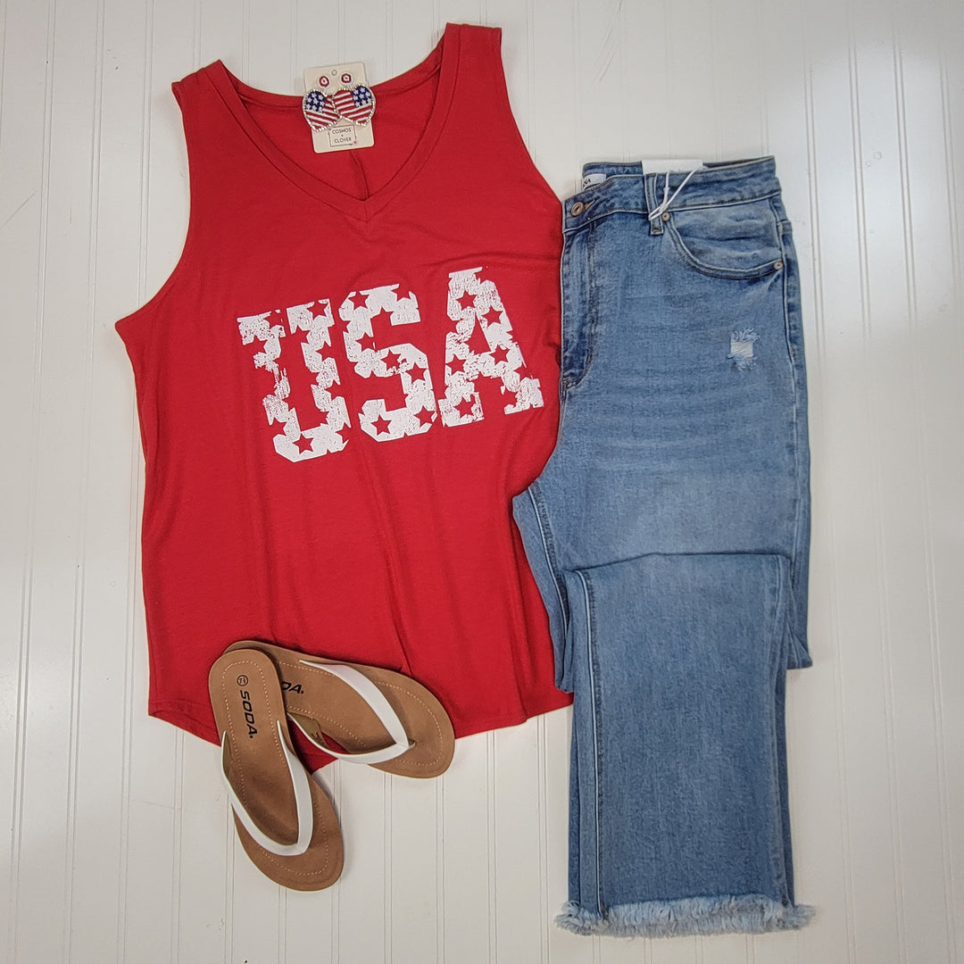 The USA Graphic V-Neck Tank by Cosmos + Clover Boutique is styled with light blue distressed jeans and tan flip-flops, displayed on a white wooden surface.
