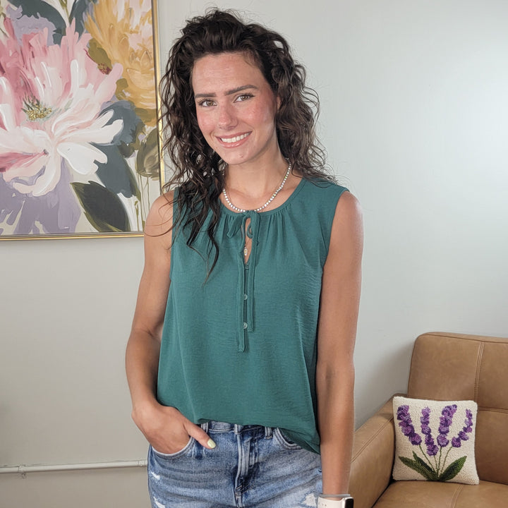 A woman with wavy brown hair smiles indoors, wearing the Eileen Sleeveless Top by Staccato and denim shorts. She accessorizes with a necklace and watch, standing before floral decor and a tan couch with a lavender pillow.