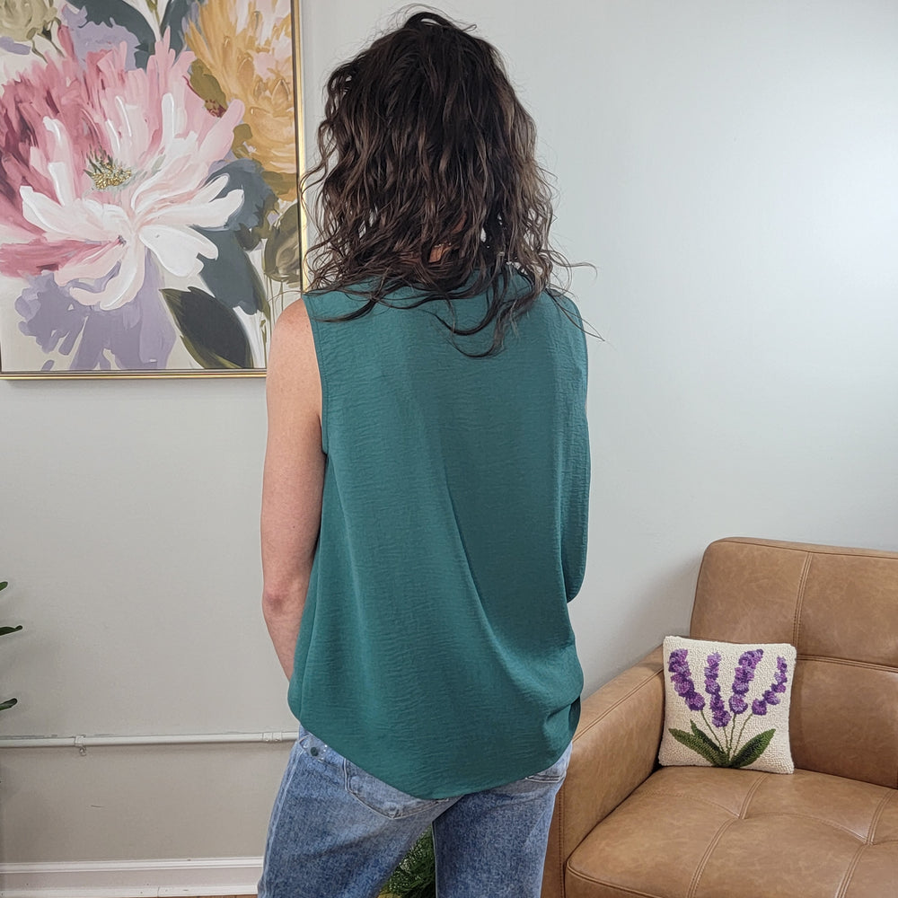 A person with wavy brown hair, wearing the Staccato Eileen Sleeveless Top in jade and jeans, stands facing away in a living room with a tan couch, floral cushion, and a large flower painting on the wall.