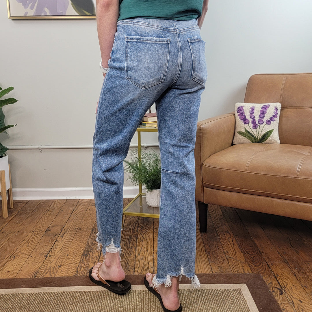 Indoors, a person wears Risen's Maya Crop Straight Jeans—light blue, high rise, with a frayed hem—and black sandals. A tan armchair with a floral pillow and houseplants appear in the background.