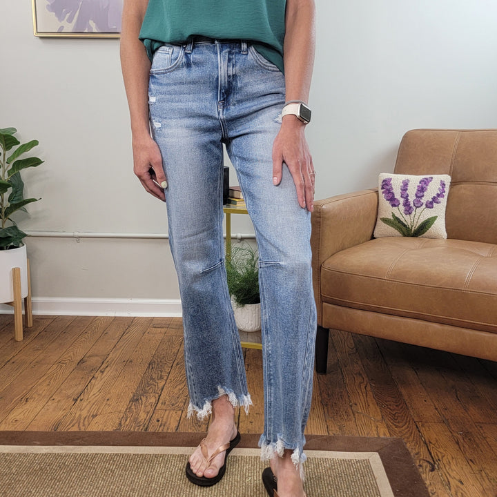 Indoors, a person wears Risen's Maya Crop Straight Jeans in light blue distressed denim with frayed hems, a green top, black sandals, and a smartwatch. Behind them are a tan chair with a lavender pillow and a green plant.