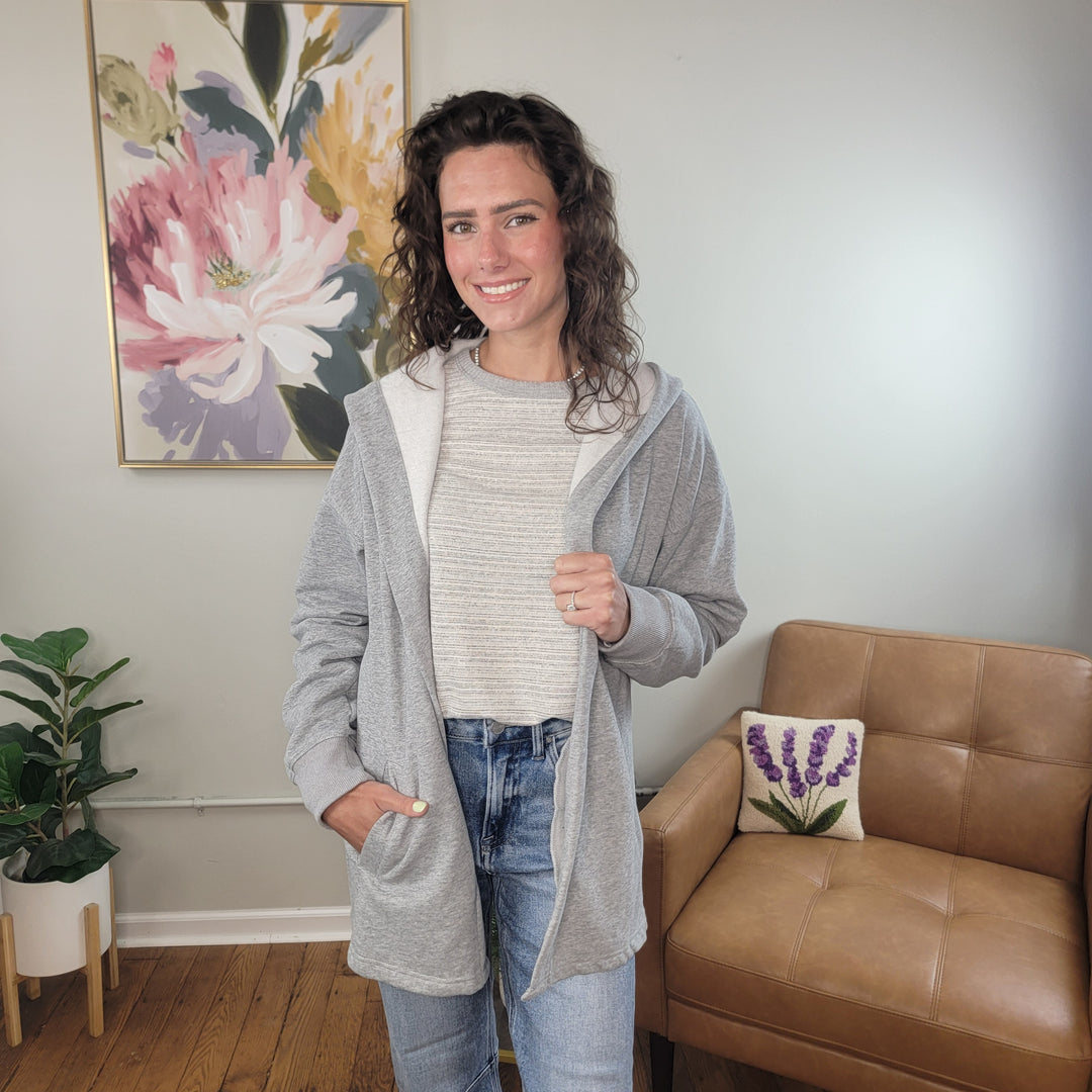 A woman with wavy brown hair smiles while wearing the Staccato Irene Cardigan over a striped shirt and jeans, standing indoors beside a tan chair and a floral painting.