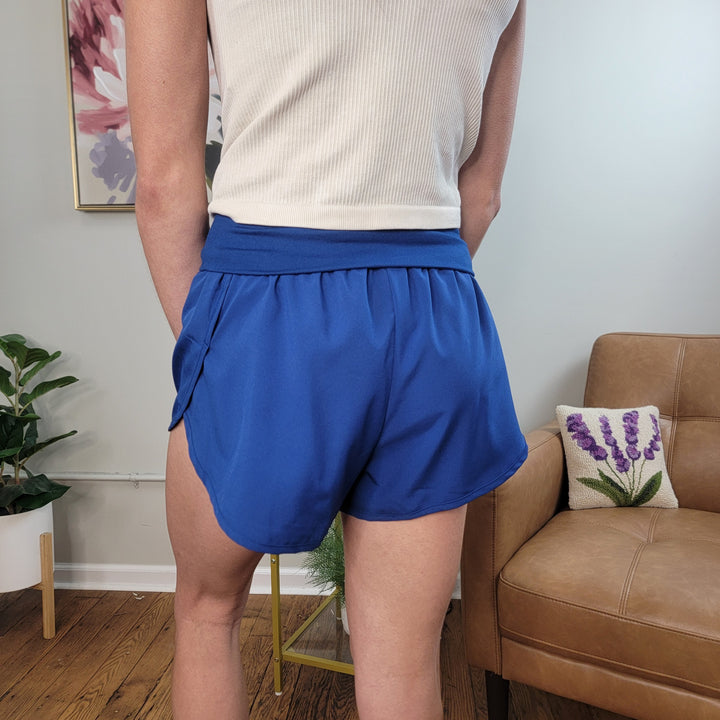 A person wearing a white tank top and ZENANA Wrenlee Running Shorts in blue stands indoors with their back to the camera. A tan chair with a lavender pillow, a plant, and a floral painting are visible in the background.