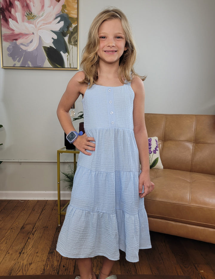 A smiling blonde girl stands indoors on a wooden floor, wearing the Cutie Patootie Girls Myla Dress and a smartwatch. A tan couch, plant, and floral painting are behind her—ideal for warm days and playful style.