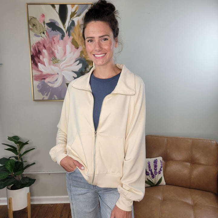 A woman with dark hair in a bun smiles in a cozy room, wearing the HYFVE, Inc Bryar Full Zip Scuba jacket over a blue shirt. A tan leather chair, potted plants, and floral art decorate the space behind her.