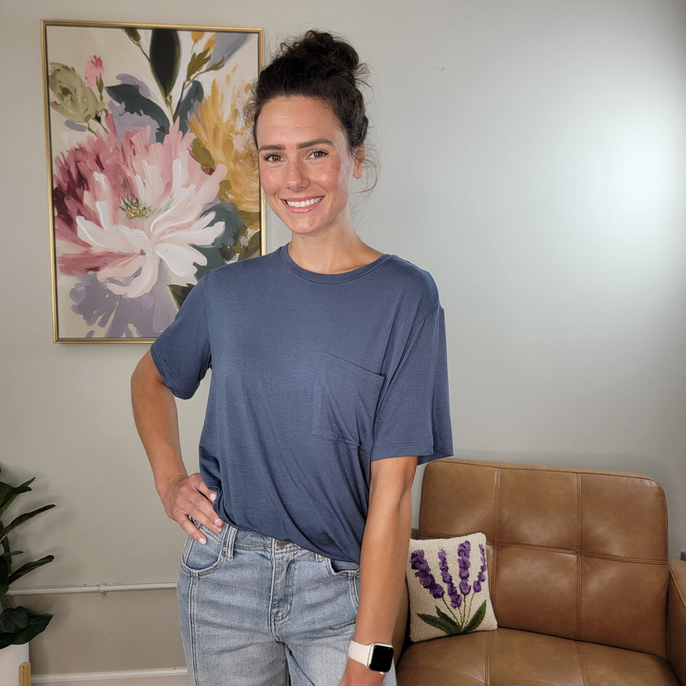 A woman with dark hair tied up smiles indoors, wearing the HYFVE, Inc Khloe Tee—a blue crew neck pocket tee in soft, silky fabric—and light jeans. A floral painting and a tan chair with a lavender pillow are in the background.