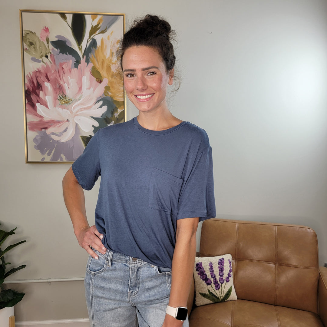 A woman with dark hair tied up smiles indoors, wearing the HYFVE, Inc Khloe Tee—a blue crew neck pocket tee in soft, silky fabric—and light jeans. A floral painting and a tan chair with a lavender pillow are in the background.