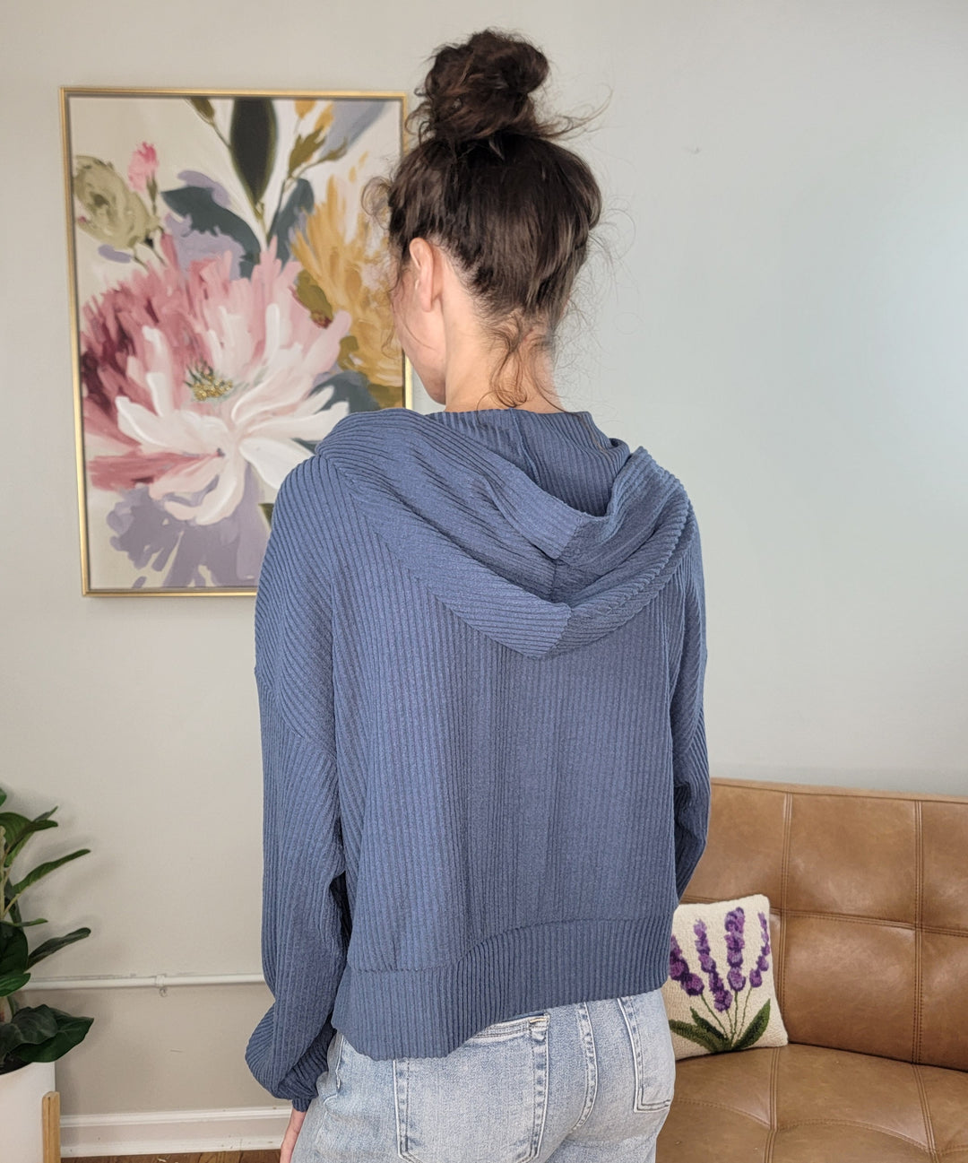 A person with brown hair in a messy bun wears the HYFVE, Inc Raylee Cropped Hoodie with an oversize fit and light jeans, standing with their back to the camera in a living room with floral decor and a tan couch.