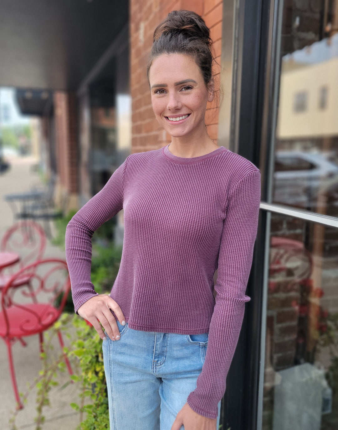 A woman with brown hair in a bun wears the HYFVE, Inc Eliza Long Sleeve in purple and light blue jeans, smiling outside near a brick wall and window, with red chairs and tables visible in the background.